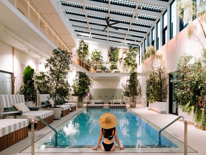 A woman sits facing an indoor pool as seen from behind wearing a hat.