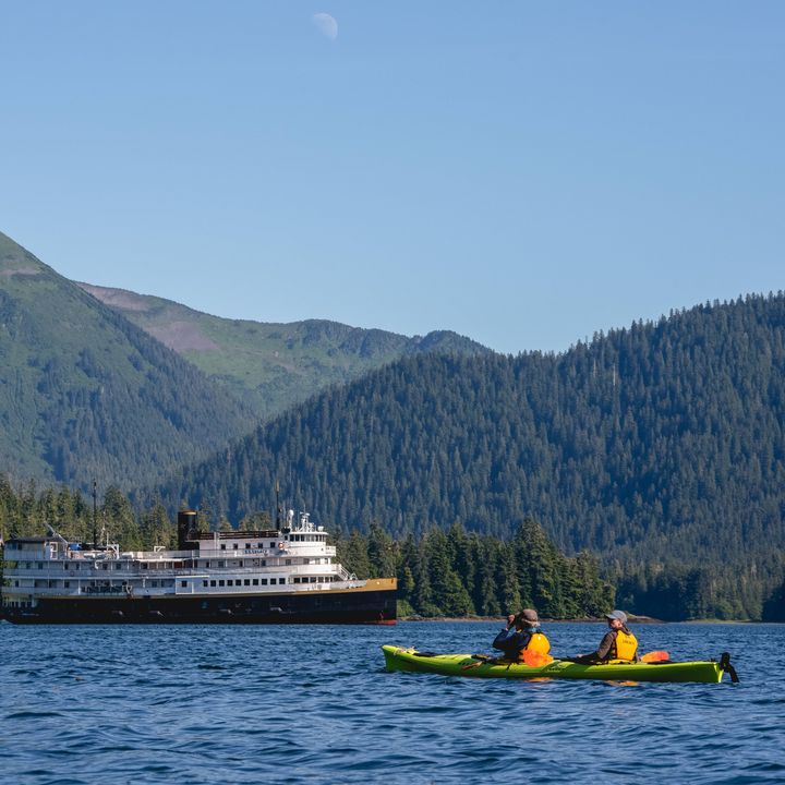 A kayak is seen on the water in front of an expedition cruise boat in a mountainous area.