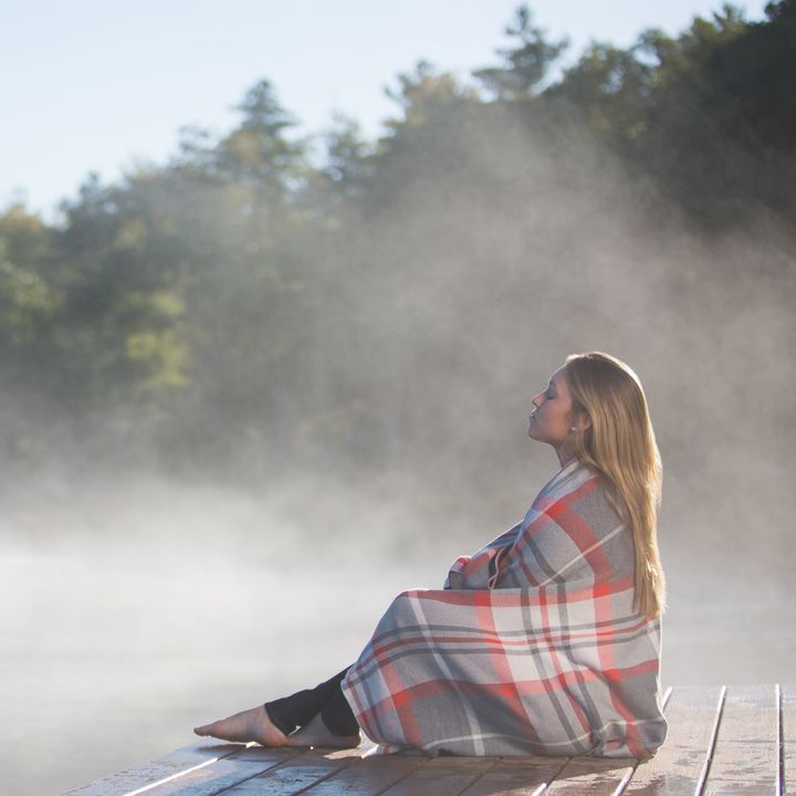 A woman sits on a dock at the water’s edge as steam rises off it, wrapped in a blanket.