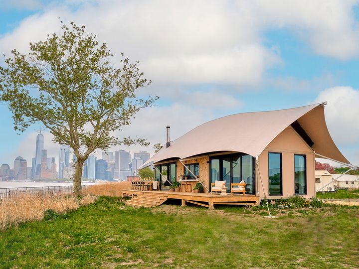 A glamping tent on a grassy area next to a tree with views of the New York City skyline behind it.