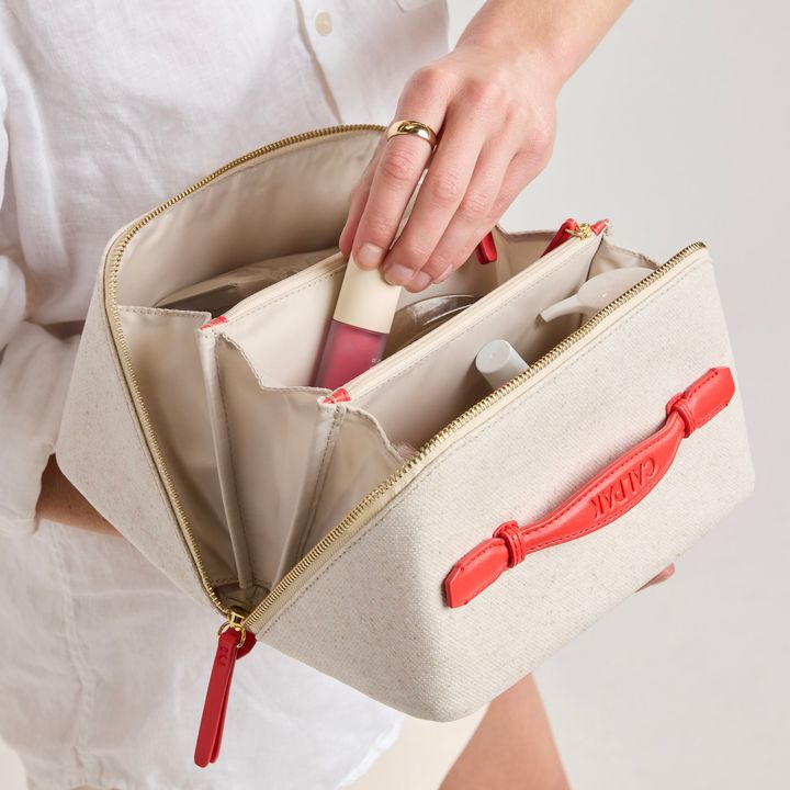 A hand reaching into the Calpak lay flat toiletry bag while open to put in lipstick.