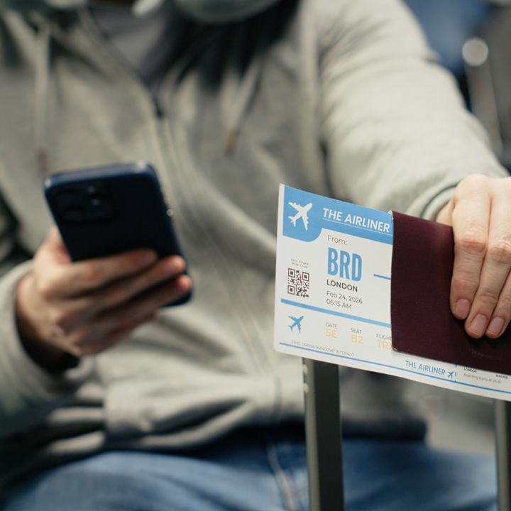 A man sitting at the airport visible from the neck down holding a phone in one hand and a passport and ticket in the other leaning on this suitcase handle.