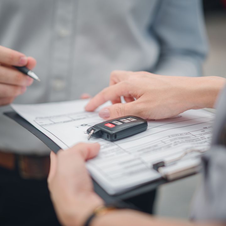 One person is seen with hands only signing a clipboard of paperwork, while another points to a place to sign with car keys on top of the papers.