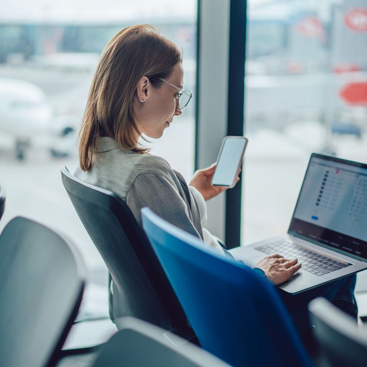 A woman sits in a chair at an airport holding her phone and looking at her laptop computer.
