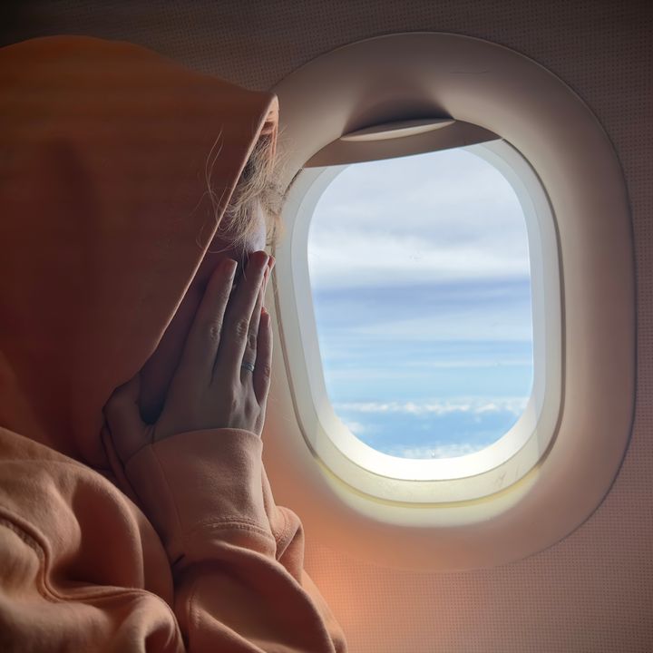 A person on an airplane next to a window covering their face with a hood on.