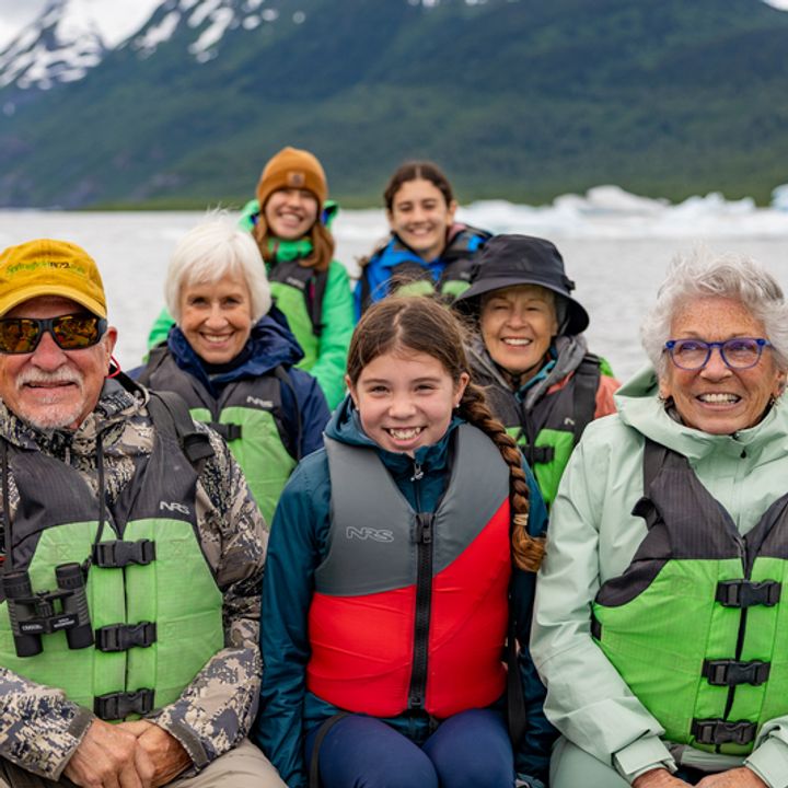 A group smiles for a photo in a boat next to snow-capped mountains with young children and senior-age grandparents.