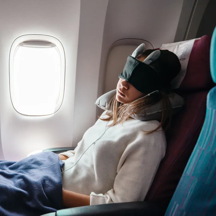A woman sleeping on a plane while wearing an eye mask next to a window.