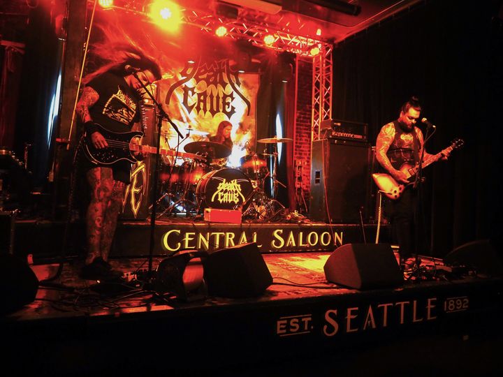 A rock band performs on a stage lit by red and orange lights with signage for Central Saloon in Seattle.