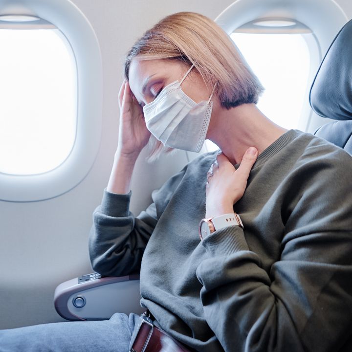 A woman is seen at a window seat on an airplane wearing a surgical face mask with one hand on her head and the other at her throat as if she’s ill.