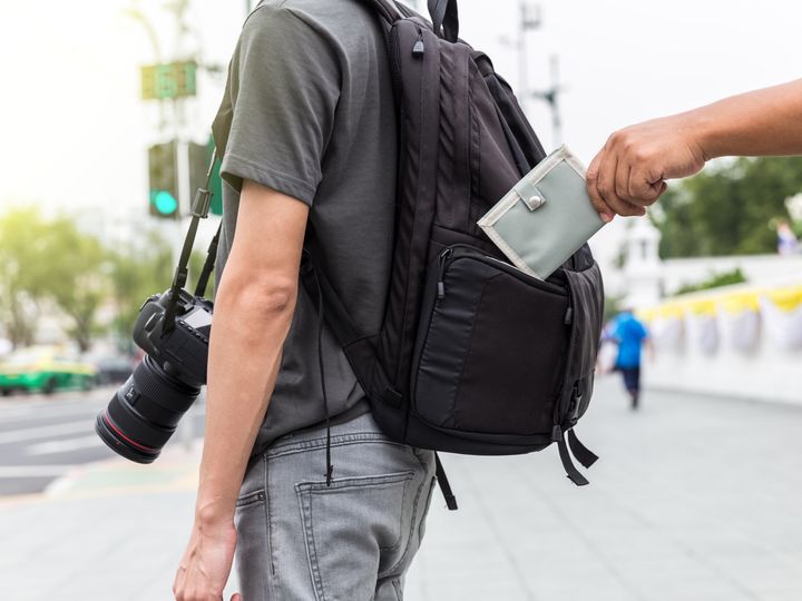 A pickpocket stealing a wallet out of the backpack of a tourist standing on the street carrying a camera.