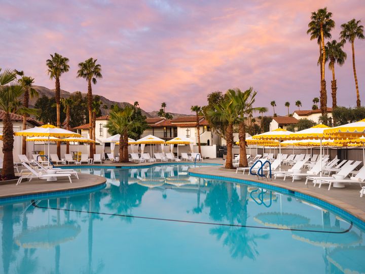 A pool surrounded with white chairs, yellow striped umbrellas, and palm trees at sunset at Omni Rancho Las Palmas Resort & Spa.