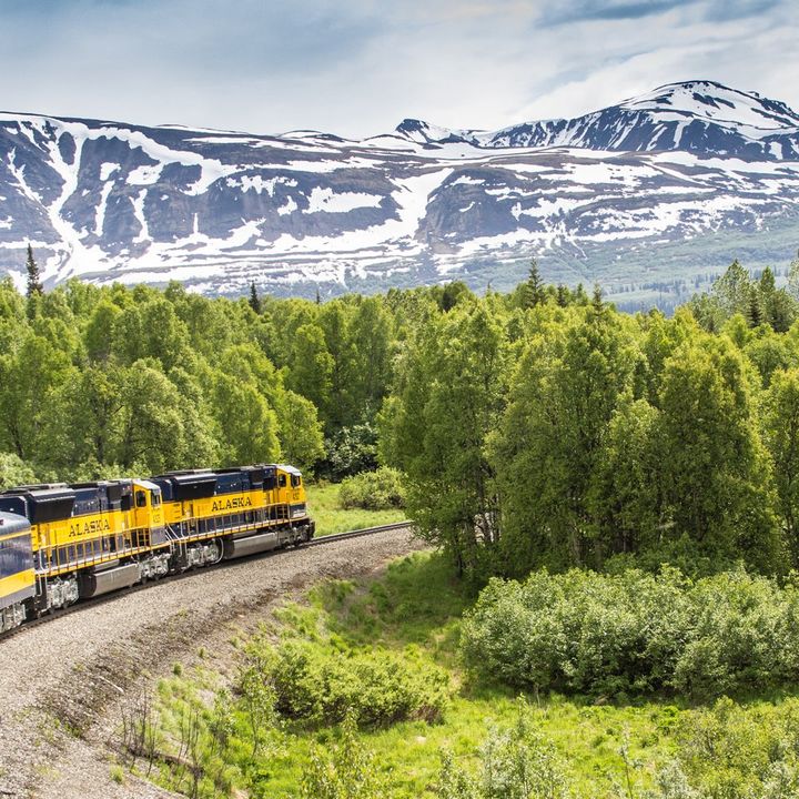 A yellow train labeled "Alaska" passes along tracks next to snow-covered mountains.