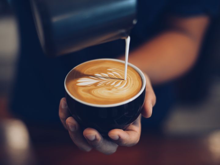 A hand is holding a cup with milk being artfully poured into a latte.