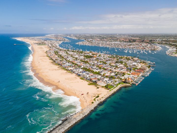An aerial view over Newport Beach, including Balboa Island and the harbor, with a wide beach area and blue-turquoise water.