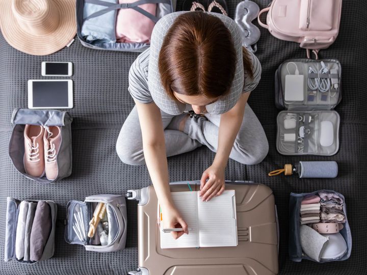 An overhead shot of a woman packing, surrounded by packing cubes and accessories of various sizes as she writes on a notebook on top of her suitcase.