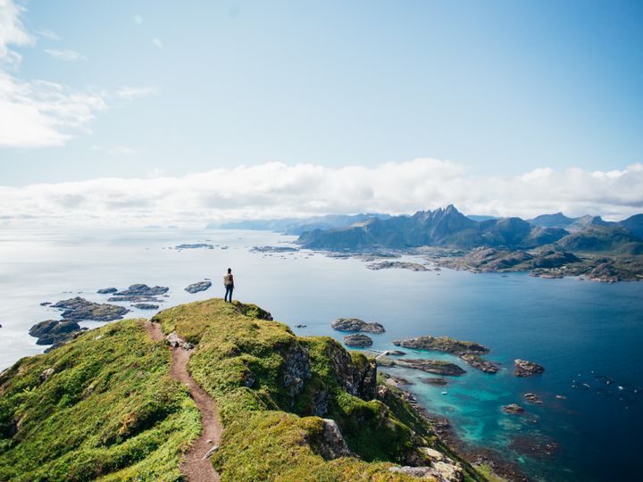 A hiker stands on top of a mountain after a hike in Lofoten Islands in Norway looking out over rocks, water, and mountains.