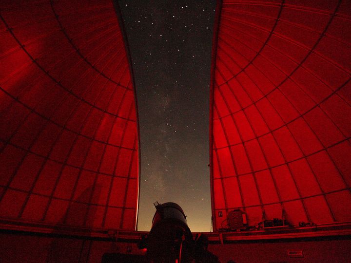 Looking out from inside an observatory with a sliver or stars and the interior lit up in red lights.