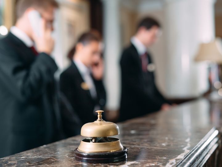 A marble hotel desk with a bell to ring for service on it, with people blurred out in the background on the phone.