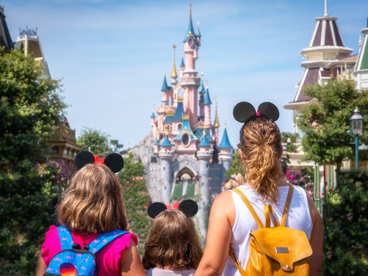 A woman and her two daughters stand in Disneyland looking at the castle, wearing backpacks.