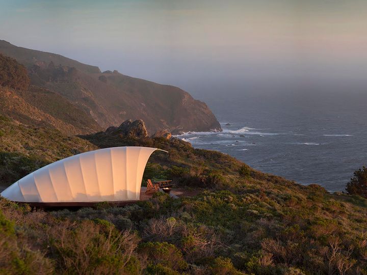 A large white tent with a deck with chairs facing the ocean surrounded by the natural beauty of Big Sur at sunset.