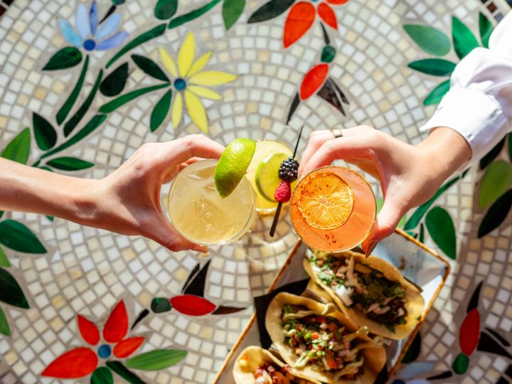 An overhead view of two sets of hands clinking cocktails over a mosaic tiled-table with tacos.