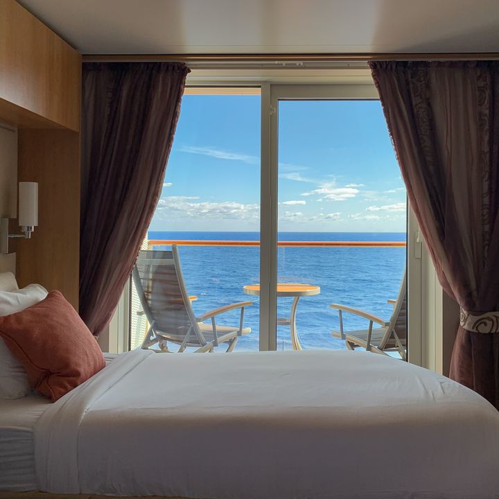 A cruise ship stateroom with a bed next to a balcony with two chairs overlooking the blue ocean.