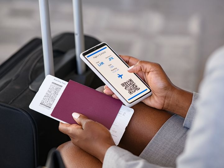 A woman’s hand is seen holding a smart phone with her digital boarding pass visible on her app, next to her passport in her other hand and luggage.