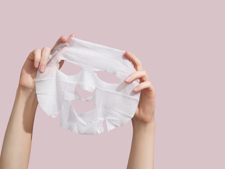 Female hands holding sheet of white mask on pink background.