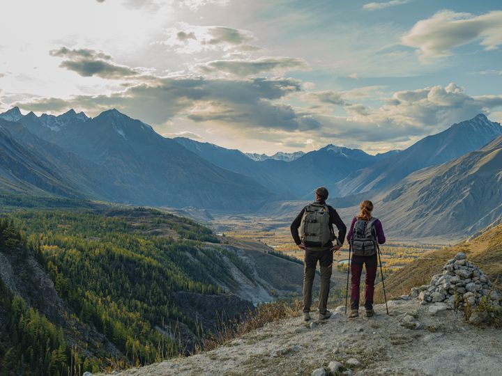 A couple of hikers with backpacks standing on mountain peak looking at vast valley, panoramic view of snow capped mountains and autumn forest in Mongolia