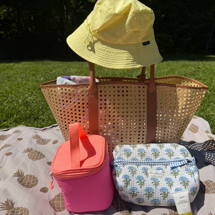 A beach bag on a towel with a hat on the handle, cooler bag, and a toiletry bag.