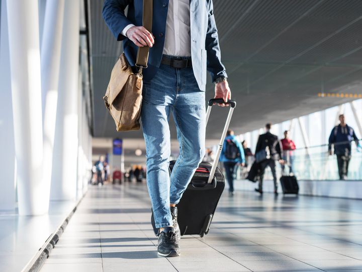 A man with a shoulder bag and hand luggage walking in airport terminal without his face visible.