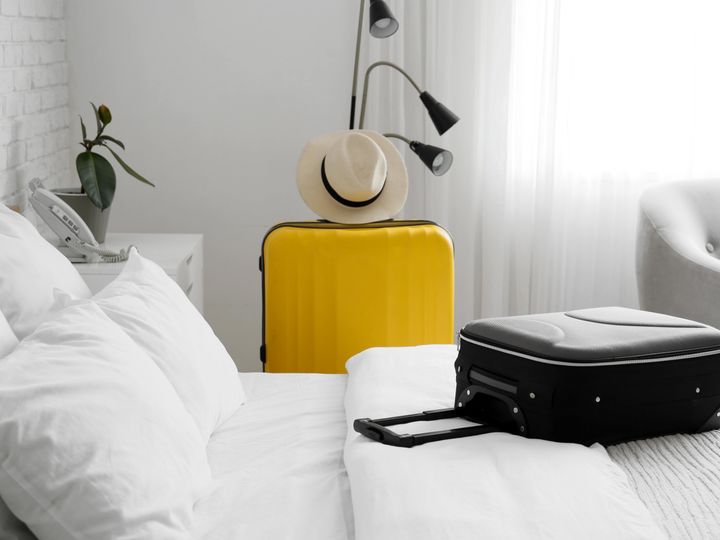 A black suitcase rests on a bed with white linens in a white room, and a standing yellow suitcase with a hat on top stands next to the bed.
