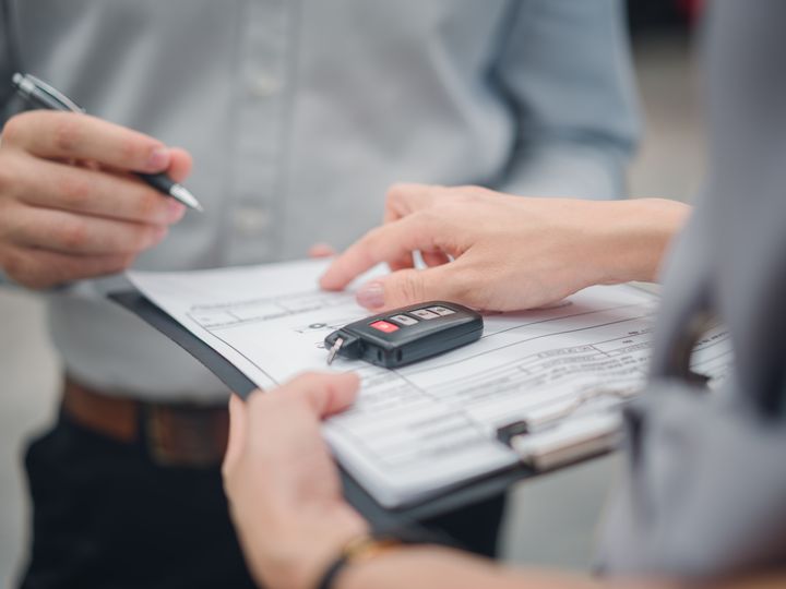 One person is seen with hands only signing a clipboard of paperwork, while another points to a place to sign with car keys on top of the papers.