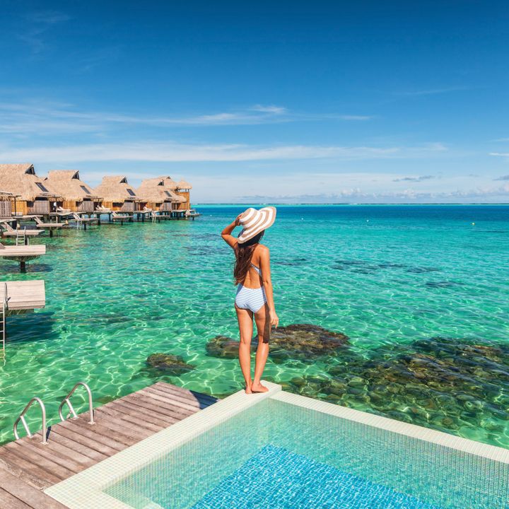 A woman standing next to a pool looking out over the ocean next to a row of overwater bungalows.