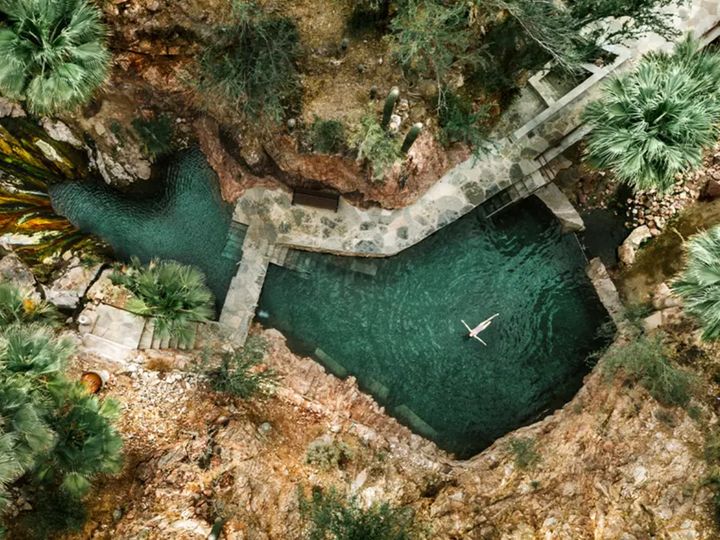 An overhead drone shot of someone swimming in a hot spring surrounded by rocky cliffs and palm trees at Castle Hot Springs.