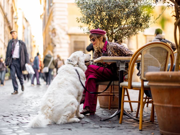 A stylish woman enjoys coffee at an outdoor cafe in Rome while bonding with her large white dog.
