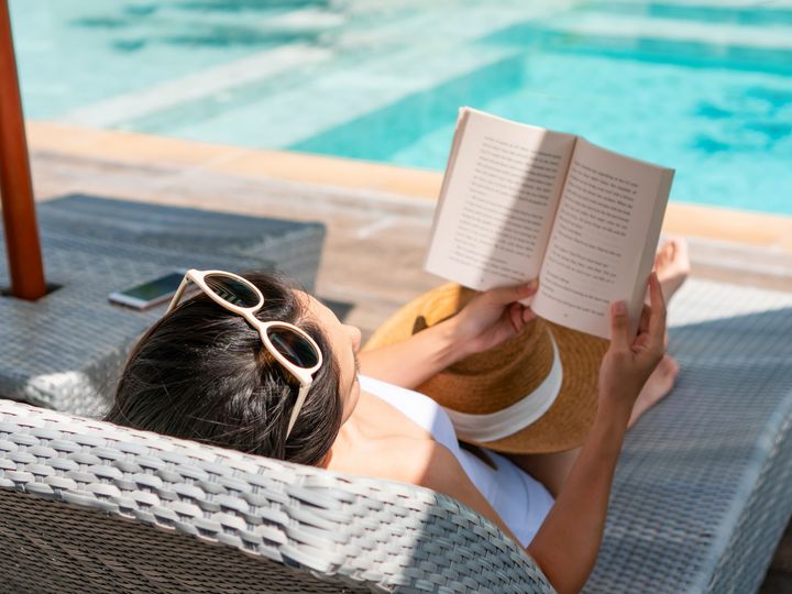 A woman is seen from behind on a pool chair reading a book by the water with her sunglasses on top of her head.
