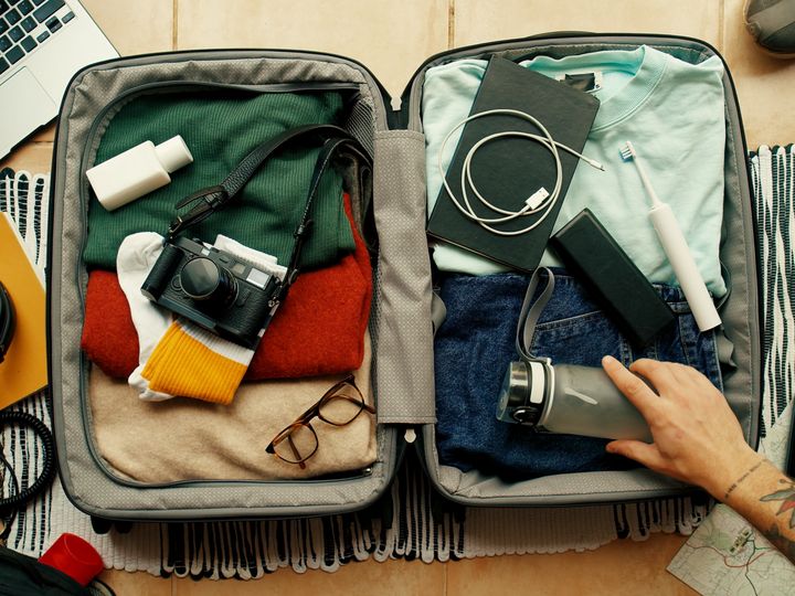 A hand is seen reaching into a suitcase packed full of clothes and accessories next to a laptop and other small items like headphones and a wallet and keyboard.
