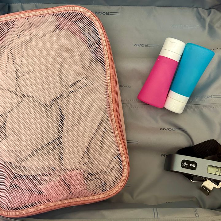 A suitcase half is filled with just a packing cube, blue and pink toiletry bottles, and a digital luggage scale.