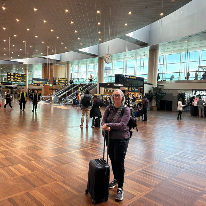 A woman stands in a crowded airport with her hands on her rolling bag wearing a long sleeve shirt and jogger pants.