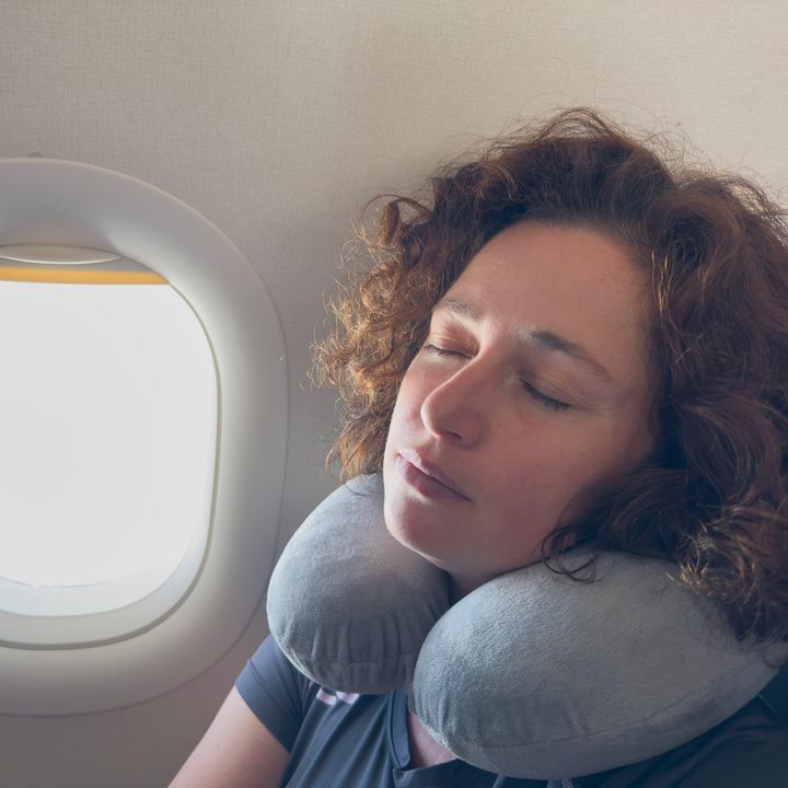A woman with curly hair sleeping next to a plane window wearing a neck pillow.