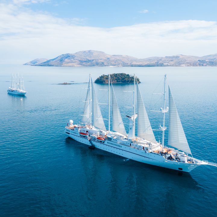 A tall sail ship from Windstar cruises is seen sailing not too far from land with a smaller ship behind it.