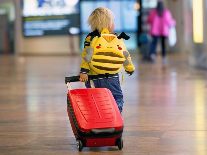 A child pulling a small red suitcase while wearing a bee backpack.