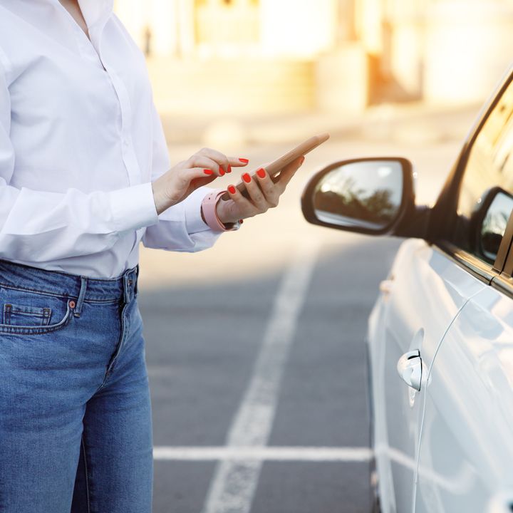A woman from the neck down is seen on her phone next to a car.