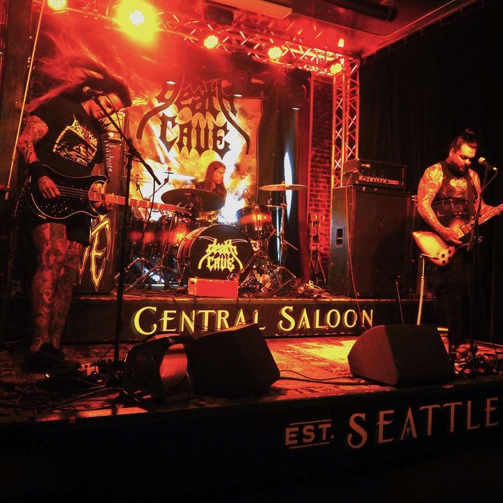 A rock band performs on a stage lit by red and orange lights with signage for Central Saloon in Seattle.