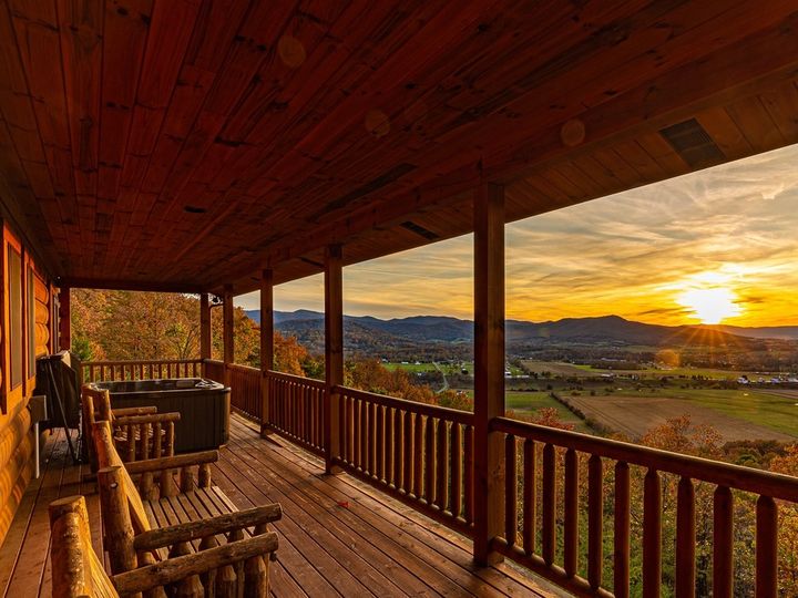 One side of a log cabin with a long wooden deck, two chairs, and views stretching over fields and mountains at sunset in fall.