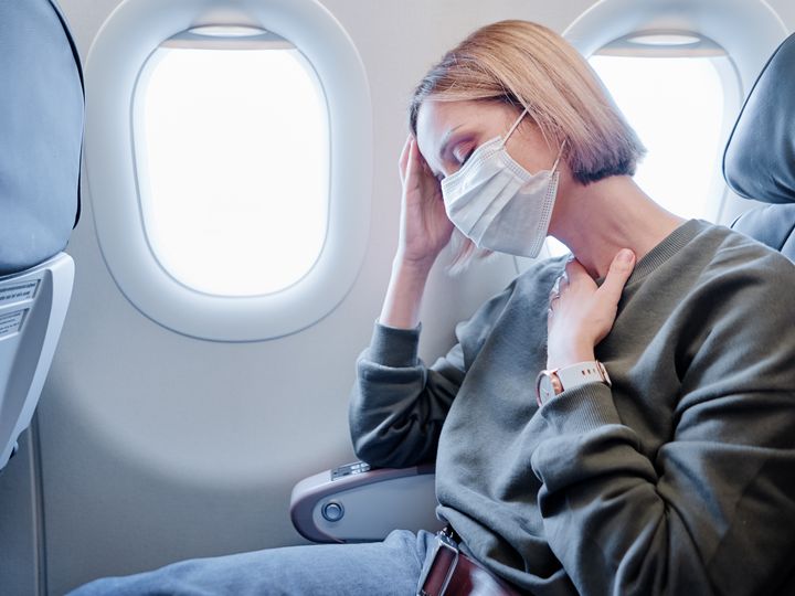 A woman is seen at a window seat on an airplane wearing a surgical face mask with one hand on her head and the other at her throat as if she’s ill.