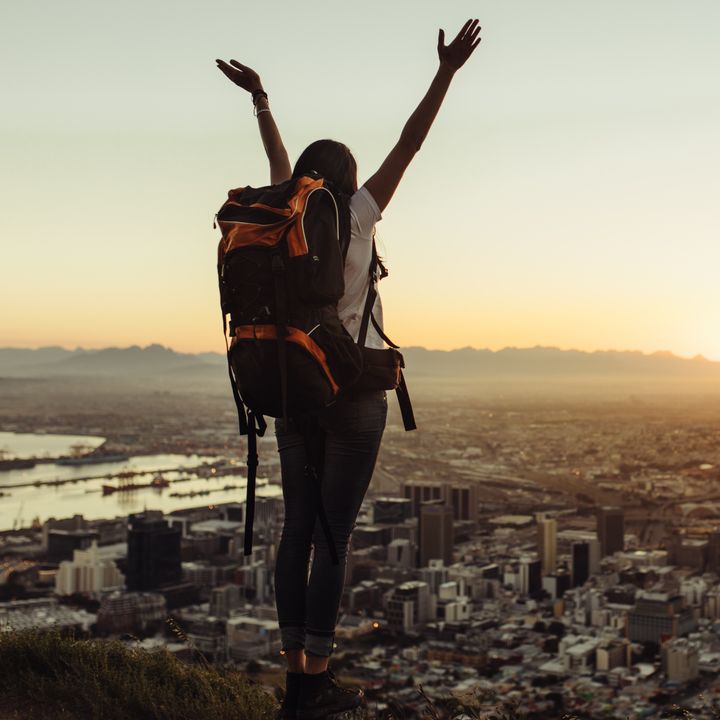 A woman standing on top of a mountain alone wearing a backpack with her hands raised in the air, looking over the city below at sunrise.