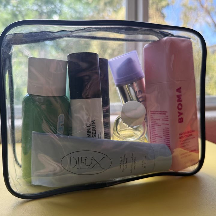 A clear Packism TSA-friendly travel bag filled with products on a table in front of a window.
