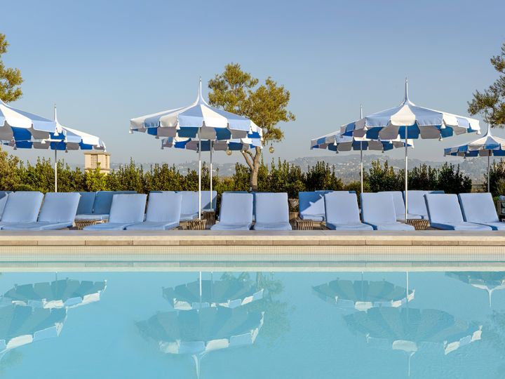 The rooftop pool at the Maybourne Beverly Hills with rows of white chairs, blue and white striped umbrellas, and LA views.